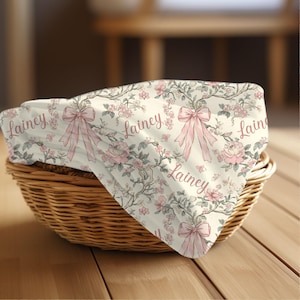 May include: A white blanket with a floral and bow pattern, featuring the name "Lainey" in pink script. The blanket is folded in a wicker basket, with a wooden surface in the foreground.