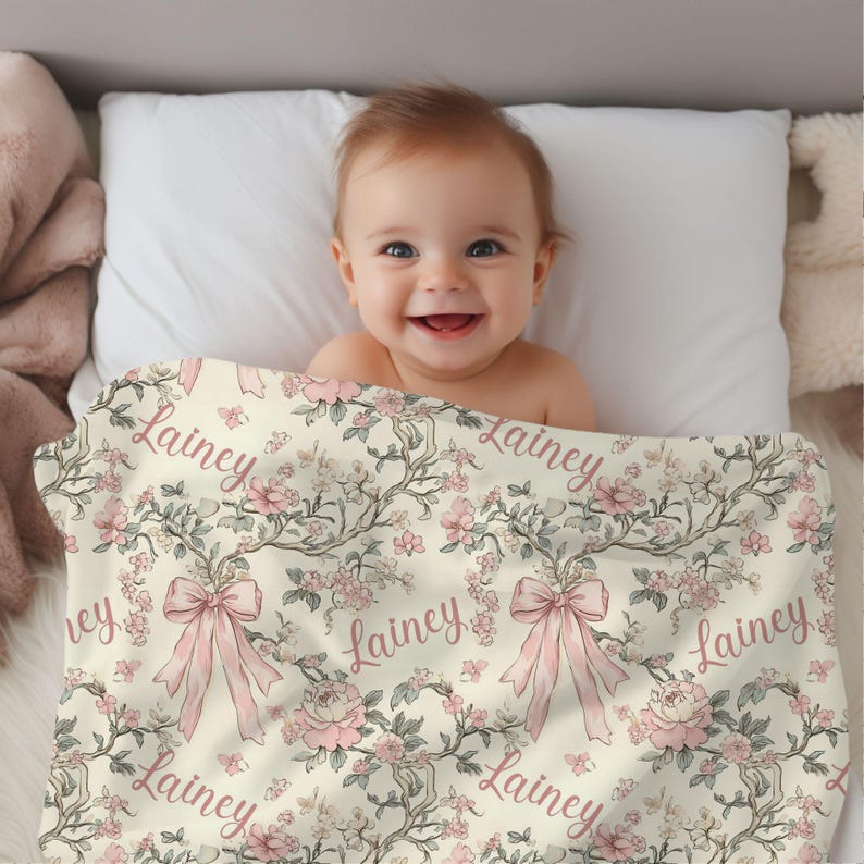 May include: A cream-colored baby blanket with a floral design and the name "Lainey" repeated across it. The blanket features pink flowers, bows, and branches. A smiling baby is lying on a white pillow, partially covered by the blanket.