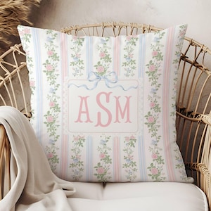May include: Square decorative pillow with a floral and striped pattern in pastel pink, blue, and white. The pillow features a monogram with the letters "ASM" in pink, and is adorned with a light blue ribbon.