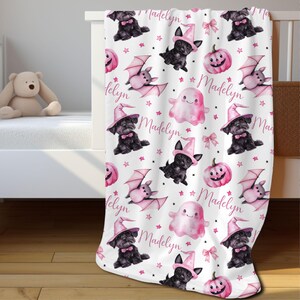 May include: Pink and white baby blanket with a Halloween theme. The blanket features black dogs wearing witch hats, bats, ghosts, pumpkins, and stars. The name "Madelyn" is repeated throughout the blanket.