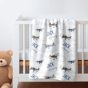 May include: White baby blanket with a blue and brown airplane pattern. The blanket has the names "Jack Henry" repeated throughout the pattern.