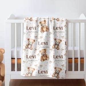 May include: A white baby blanket with a brown teddy bear pattern. The blanket has a repeating pattern of brown teddy bears with bows, brown polka dots, and the name "Levi Jameson" written in brown cursive.