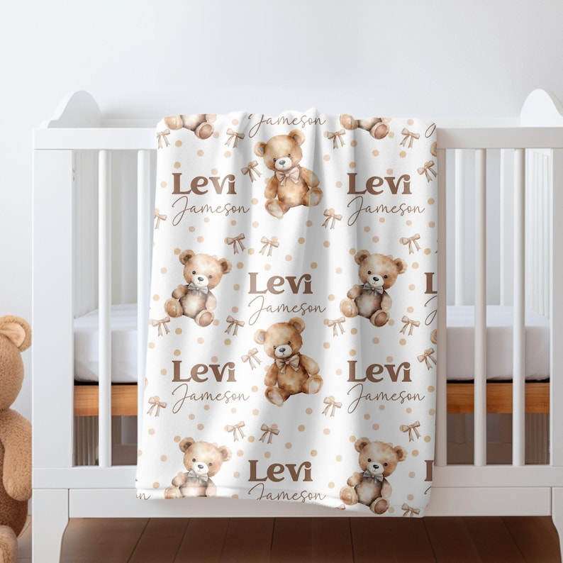 May include: A white baby blanket with a brown teddy bear pattern. The blanket has a repeating pattern of brown teddy bears with bows, brown polka dots, and the name "Levi Jameson" written in brown cursive.