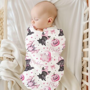 May include: A white swaddle blanket with a pink and black Halloween print. The print features bats, ghosts, pumpkins, and black cats wearing witch hats. The name "Madelyn" is repeated on the blanket.
