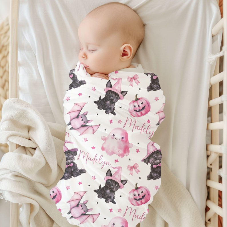 May include: A white swaddle blanket with a pink and black Halloween print. The print features bats, ghosts, pumpkins, and black cats wearing witch hats. The name "Madelyn" is repeated on the blanket.
