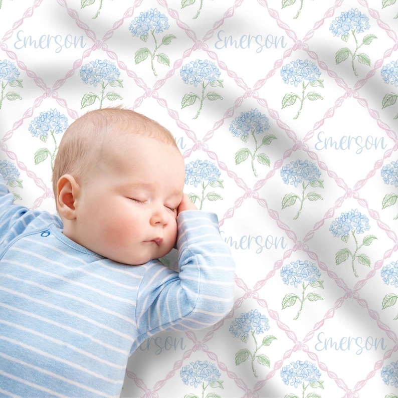 May include: A baby sleeping on a white and pink crib sheet with a blue and green floral pattern. The sheet has the name "Emerson" repeated throughout the design.