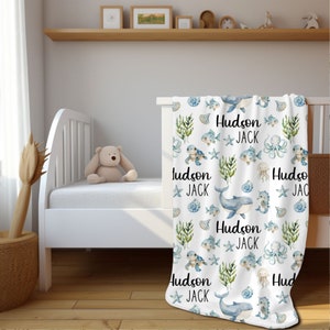 May include: White baby blanket with a blue and green watercolor pattern of sea creatures, including whales, fish, seahorses, and jellyfish. The blanket has the names "Hudson" and "Jack" repeated throughout the pattern.