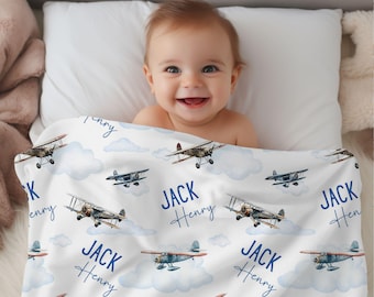 Puede incluir: Una manta de bebé blanca con un patrón de avión azul. La manta tiene el nombre "Jack Henry" repetido en todo el diseño. Un bebé está acostado en la manta, sonriendo.