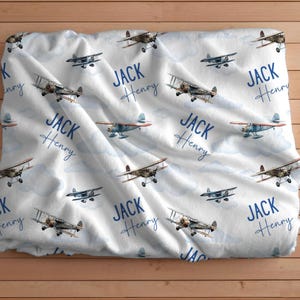 May include: White baby blanket with a blue airplane pattern. The blanket has the name "JACK HENRY" repeated throughout the design.