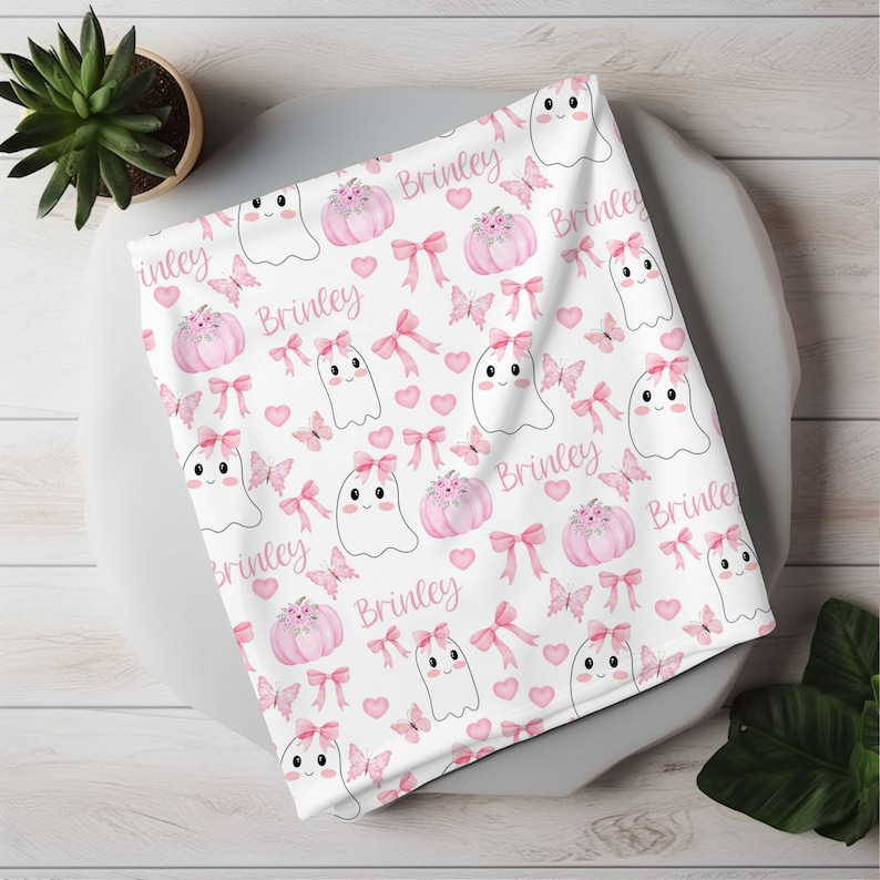 May include: A white baby blanket with a pink and white pattern of ghosts, pumpkins, butterflies, and bows. The name "Brinkley" is repeated throughout the pattern.