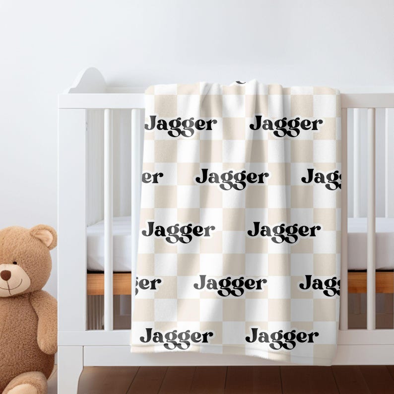 K&ouml;nnte beinhalten: Eine wei&szlig;e und beige karierte Babydecke mit dem Namen "Jagger", der in einer schwarzen Retro-Schrift wiederholt wird.