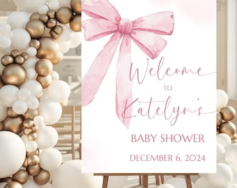 May include: A rectangular sign featuring a large watercolor-style pink bow at the top. The text reads Welcome to Katelyn's BABY SHOWER DECEMBER 6, 2024. The sign is displayed on a wooden easel surrounded by white and metallic gold balloons.