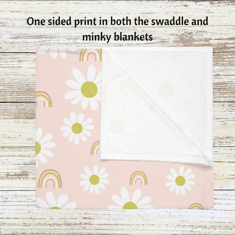 May include: A pink swaddle blanket with a white daisy and rainbow pattern. One side of the blanket is white.
