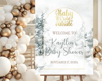 May include: A white and gold baby shower sign with a watercolor winter scene. The text reads "Baby it's cold outside" and "Welcome to Kaytlin's Baby Shower" with the date "November 17, 2024".