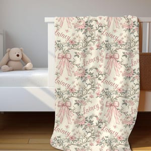 May include: A cream-colored baby blanket with a floral pattern of pink roses, bows, and the name "Lainey" in a cursive font. The blanket is draped over a white crib, with a stuffed teddy bear visible.