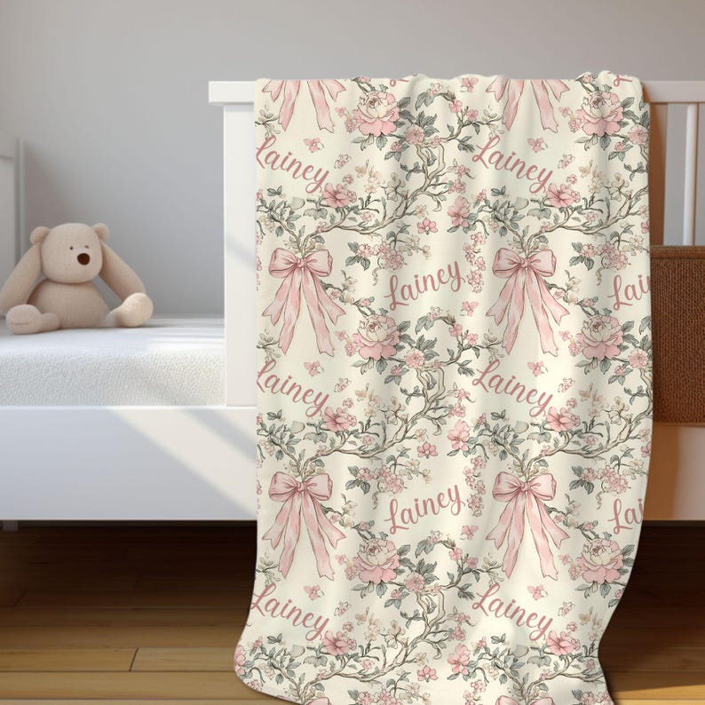 May include: A cream-colored baby blanket with a floral pattern of pink roses, bows, and the name "Lainey" in a cursive font. The blanket is draped over a white crib, with a stuffed teddy bear visible.