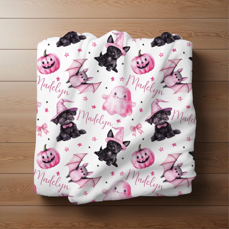 May include: A white fabric with a pink and black Halloween themed pattern. The pattern includes bats, ghosts, pumpkins, and black dogs wearing witch hats. The name "Madelyn" is repeated throughout the fabric.