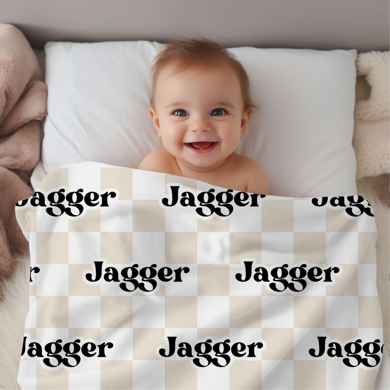 K&ouml;nnte beinhalten: Eine beige und wei&szlig;e karierte Babydecke mit dem Namen "Jagger" in Schwarz darauf gedruckt. Ein Baby liegt auf der Decke.