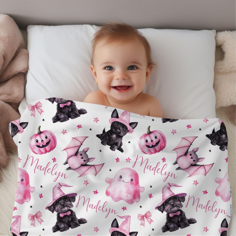 May include: A white baby blanket with a pink and black Halloween theme. The blanket features black dogs wearing witch hats, pink bats, pink ghosts, and pink pumpkins with jack-o-lantern faces. The name "Madelyn" is repeated throughout the blanket.