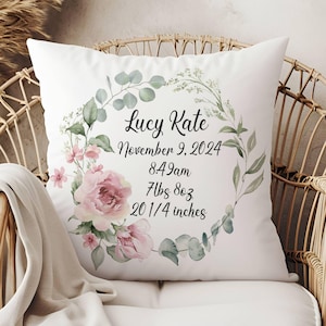 May include: White pillow with a floral wreath design. The wreath is made of pink roses, green leaves, and white flowers. The pillow has the text "Lucy Kate, November 9, 2024, 8:49am, 7lbs 8oz, 20 1/4 inches" printed in black.