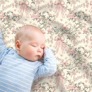 May include: A sleeping baby wearing a blue and white striped onesie rests on a floral patterned blanket. The blanket features pink bows, roses, and the name "Lainey" in a delicate script, set against a cream background.