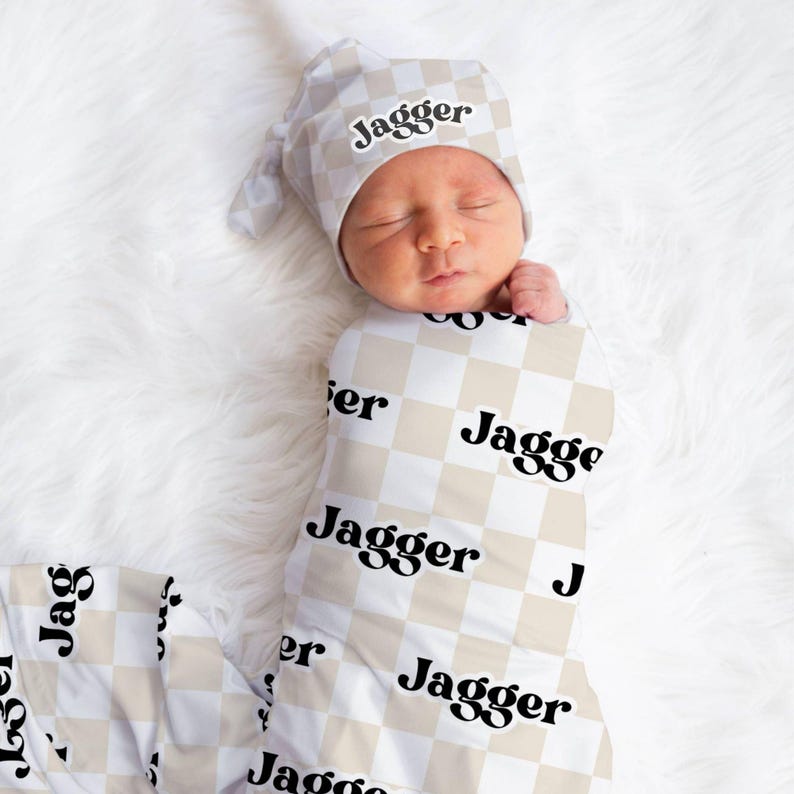 K&ouml;nnte beinhalten: Ein Baby, das in eine wei&szlig;e und beige karierte Swaddle gewickelt ist, mit dem Namen "Jagger" in Schwarz bedruckt. Das Baby tr&auml;gt eine passende M&uuml;tze mit demselben Namen.