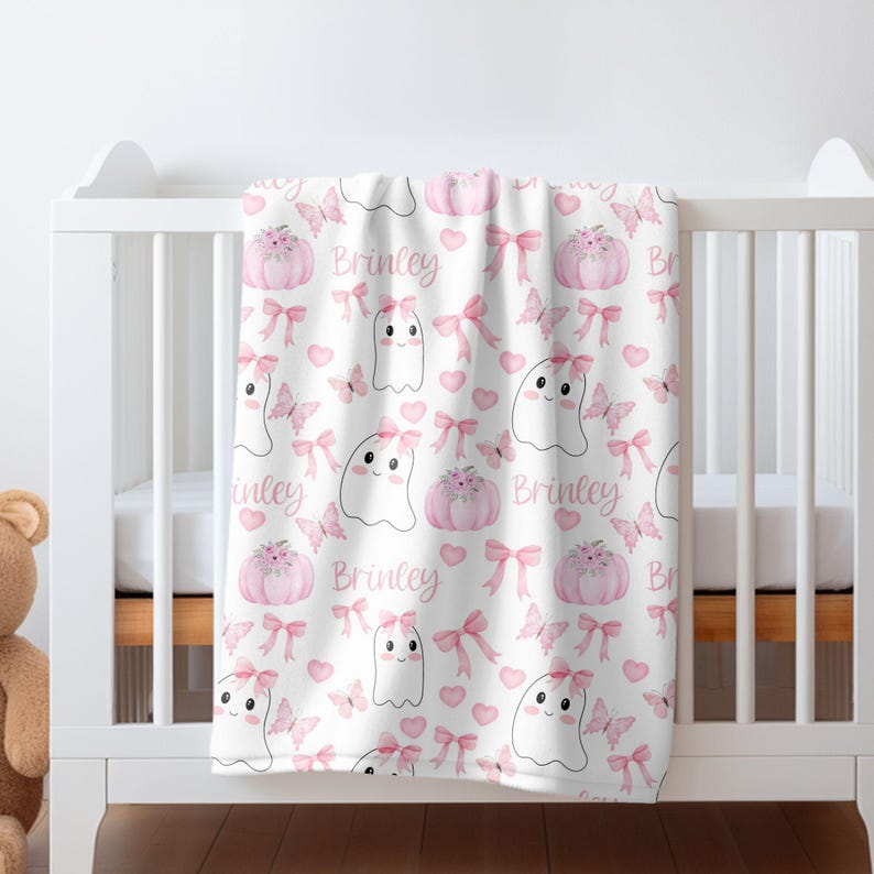 May include: A pink and white baby blanket with a repeating pattern of ghosts, pumpkins, butterflies, and bows. The blanket is personalised with the name "Brinkley".