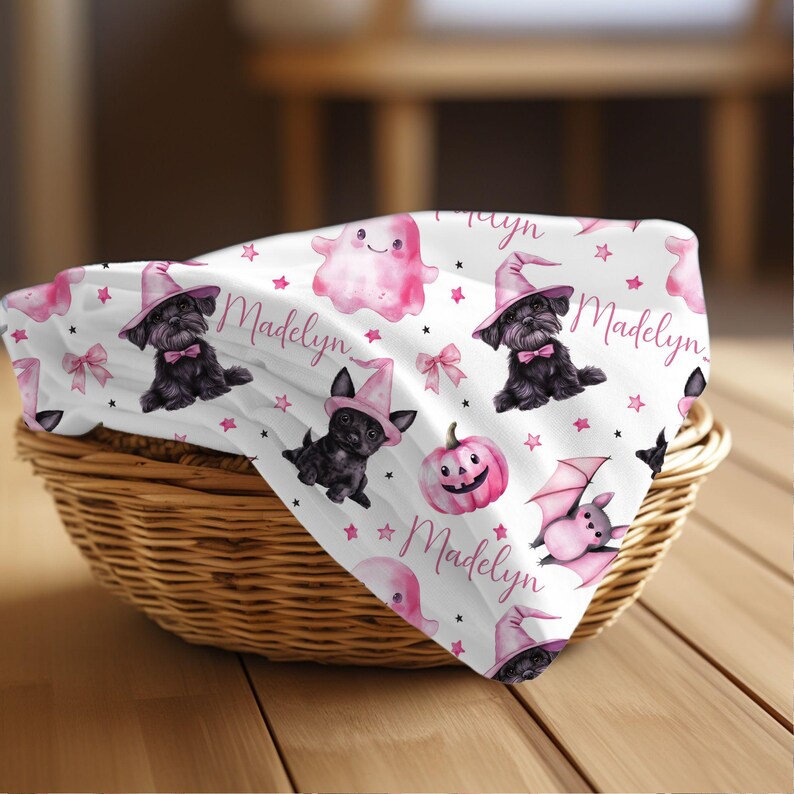 May include: A white fabric with a pink and black Halloween pattern. The pattern includes a black dog wearing a witch's hat, a ghost, a pumpkin, a bat, and pink stars. The fabric is personalized with the name "Madelyn".