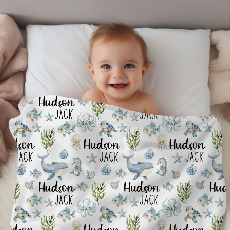 May include: A white baby blanket with a blue and green ocean-themed pattern. The blanket features sea turtles, fish, starfish, seahorses, and an octopus. The text "Hudson Jack" is repeated throughout the blanket.