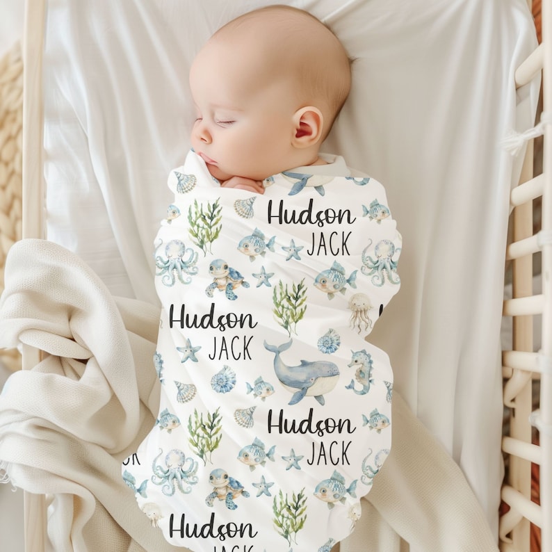 May include: A white swaddle blanket with a blue and green ocean-themed pattern. The blanket features sea creatures like whales, jellyfish, and sea turtles. The text "Hudson Jack" is repeated throughout the blanket.