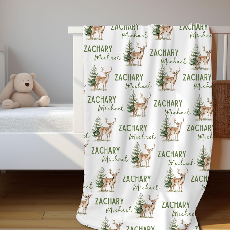 May include: White baby blanket with a repeating pattern of green trees and brown deer. The blanket has the names "Zachary Michael" printed on it.