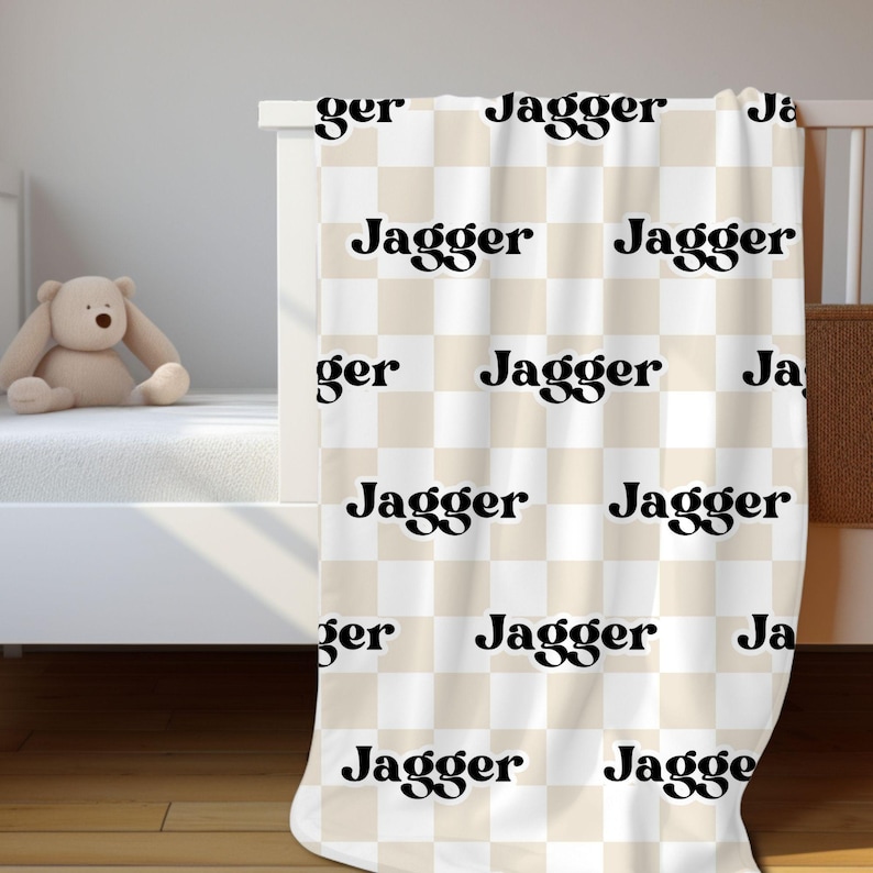 K&ouml;nnte beinhalten: Eine beige und wei&szlig;e karierte Babydecke mit dem Namen "Jagger", der in schwarzen Retro-Lettern wiederholt wird.