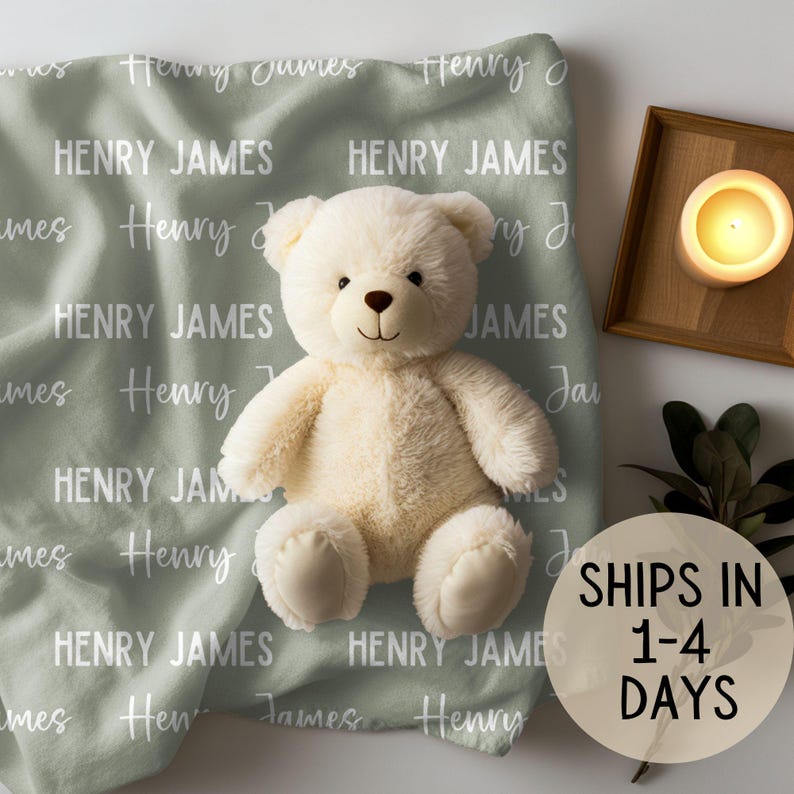 May include: A sage green baby blanket with the names "Henry James" repeated in white script. A white teddy bear sits on the blanket.