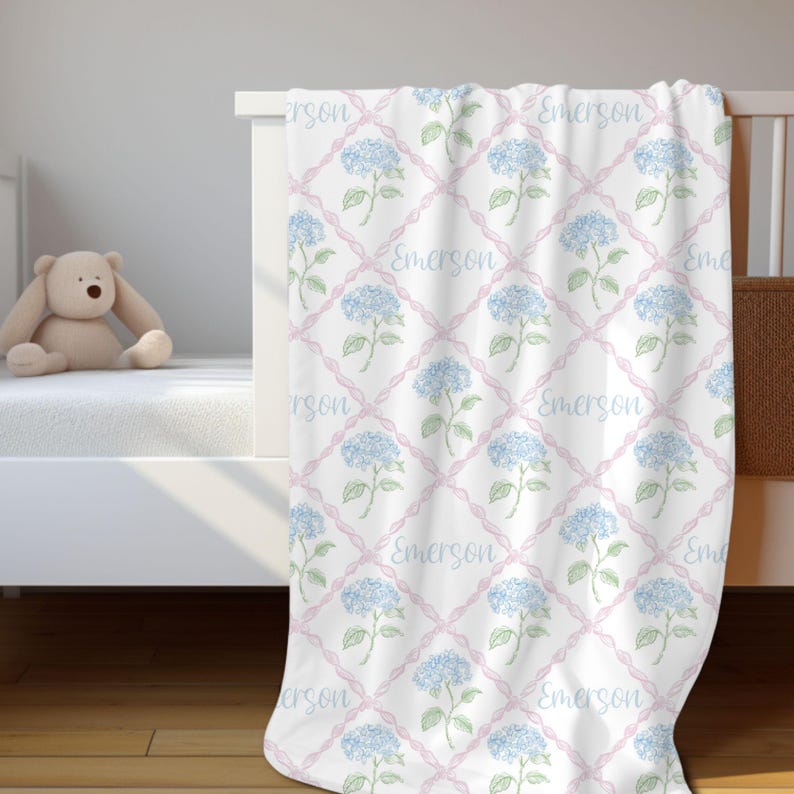 May include: A white baby blanket with a pink and blue floral pattern. The blanket features a repeating pattern of blue hydrangeas and pink ribbons with the name "Emerson" written on each ribbon.
