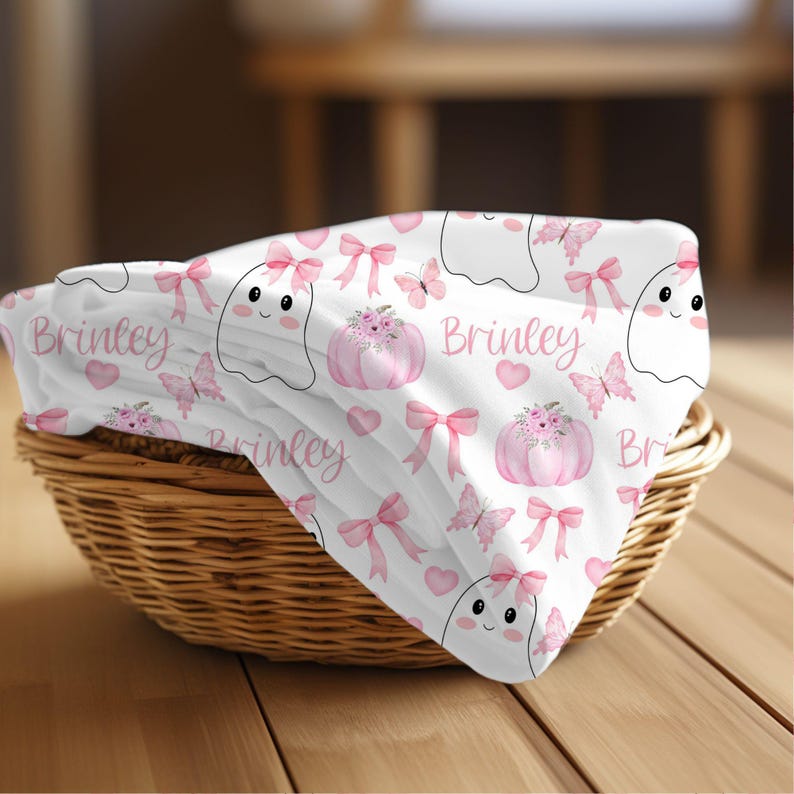 May include: A white baby blanket with a pink and white pattern of ghosts, pumpkins, butterflies, and bows. The blanket has the name "Brinkley" printed on it.