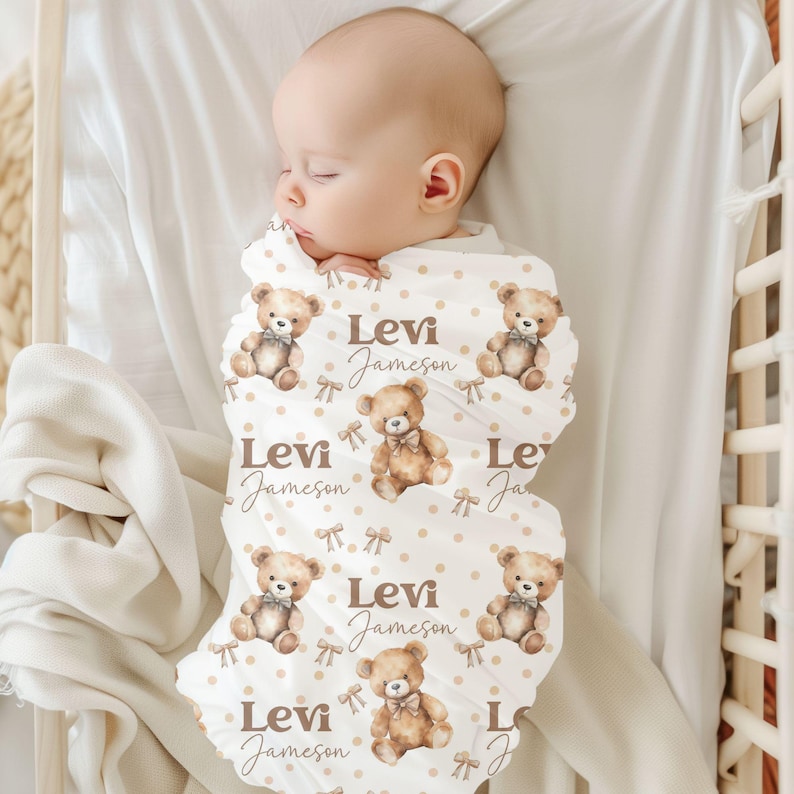 May include: A white swaddle blanket featuring a brown teddy bear design and the name "Levi Jameson" repeated throughout. The blanket is wrapped around a baby sleeping on a white cot mattress.