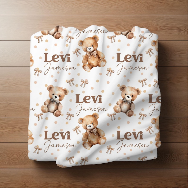 May include: White baby blanket with a repeating pattern of brown teddy bears wearing brown bows, brown polka dots, and the text "Levi Jameson" in brown.