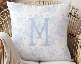 May include: Square cushion featuring a light blue floral design and a large, light blue letter "M". The cushion is placed on a white seat, with a wicker chair in the background.