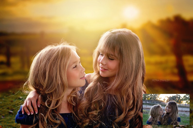May include: Two young girls with long blonde hair are looking at each other. The girl on the left has her arm around the girl on the right. They are both wearing blue dresses. The background is a blurred image of a sunset.