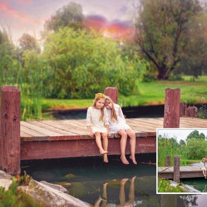 May include: Two girls wearing white dresses sit on a wooden dock over a pond. The girls are smiling and looking at the camera. The dock is surrounded by green grass and trees. The sky is a soft pink and orange.