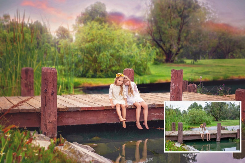 May include: Two girls wearing white dresses sit on a wooden dock over a pond. The girls are smiling and looking at the camera. The dock is surrounded by green grass and trees. The sky is a soft pink and orange.