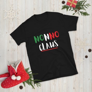 May include: Black t-shirt with the text "NONNO CLAUS" in white, green, and red. The design includes a red and green line under the word "CLAUS". The shirt is displayed on a wooden surface with Christmas decorations.
