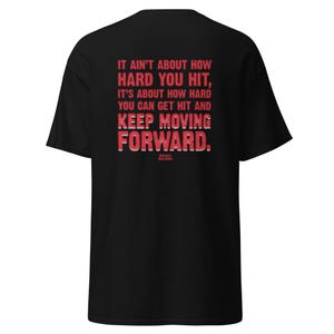 May include: Black t-shirt with the quote "IT AIN'T ABOUT HOW HARD YOU HIT, IT'S ABOUT HOW HARD YOU CAN GET HIT AND KEEP MOVING FORWARD." in red text. The text is in a bold, blocky font. The shirt is a classic crew neck.