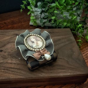 May include: A rectangular case with a brown and black checkered pattern, topped with a black and white houndstooth fabric ruffle. A circular cameo featuring a portrait is bordered by small white beads, accented with a tan bow and a pearl drop.