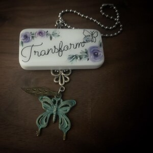 May include: A white rectangular pendant with the word "Transform" in cursive, adorned with purple floral and butterfly accents. A teal butterfly charm and a decorative metal piece hang below, suspended by a silver chain.
