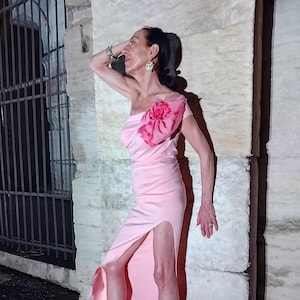 May include: A light pink, one-shoulder gown with a high slit. The dress features a pink floral accent on the shoulder. The model is wearing matching pink heeled sandals. The background is a stone wall.