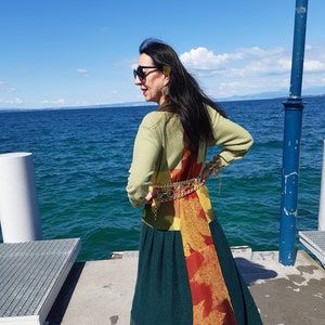 Peut inclure: Une femme porte un pull vert, une longue jupe verte avec un motif de feuilles orange et rouge, et une ceinture dorée. Elle porte des lunettes de soleil et se tient sur une jetée avec l'océan en arrière-plan.