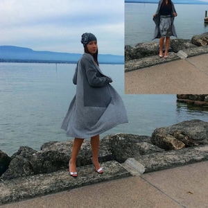 May include: A gray coat with a large patch pocket and a matching hat with a floral accent. The coat is knee-length and worn with silver heels. The background features a body of water and a cloudy sky.