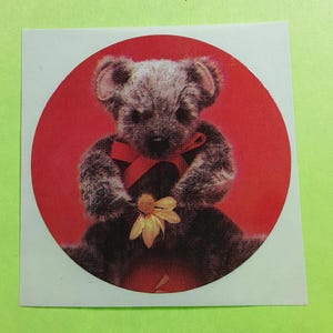 May include: A sticker featuring a teddy bear illustration. The bear is brown and gray, with a red bow and a yellow flower. The bear is set against a red circular background.