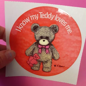 May include: A round sticker with a red background and the text "I know my Teddy loves me." A brown teddy bear with a pink bow and a smaller pink teddy bear is in the center.